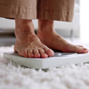 Pés de um idoso se pesando em casa, sobre uma balança, representando o cuidado e a prevenção da obesidade na terceira idade.