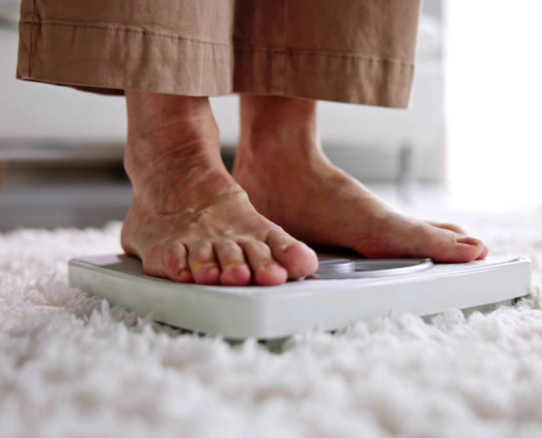Pés de um idoso se pesando em casa, sobre uma balança, representando o cuidado e a prevenção da obesidade na terceira idade.