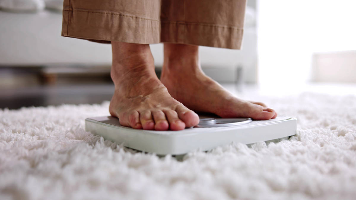 Pés de um idoso se pesando em casa, sobre uma balança, representando o cuidado e a prevenção da obesidade na terceira idade.